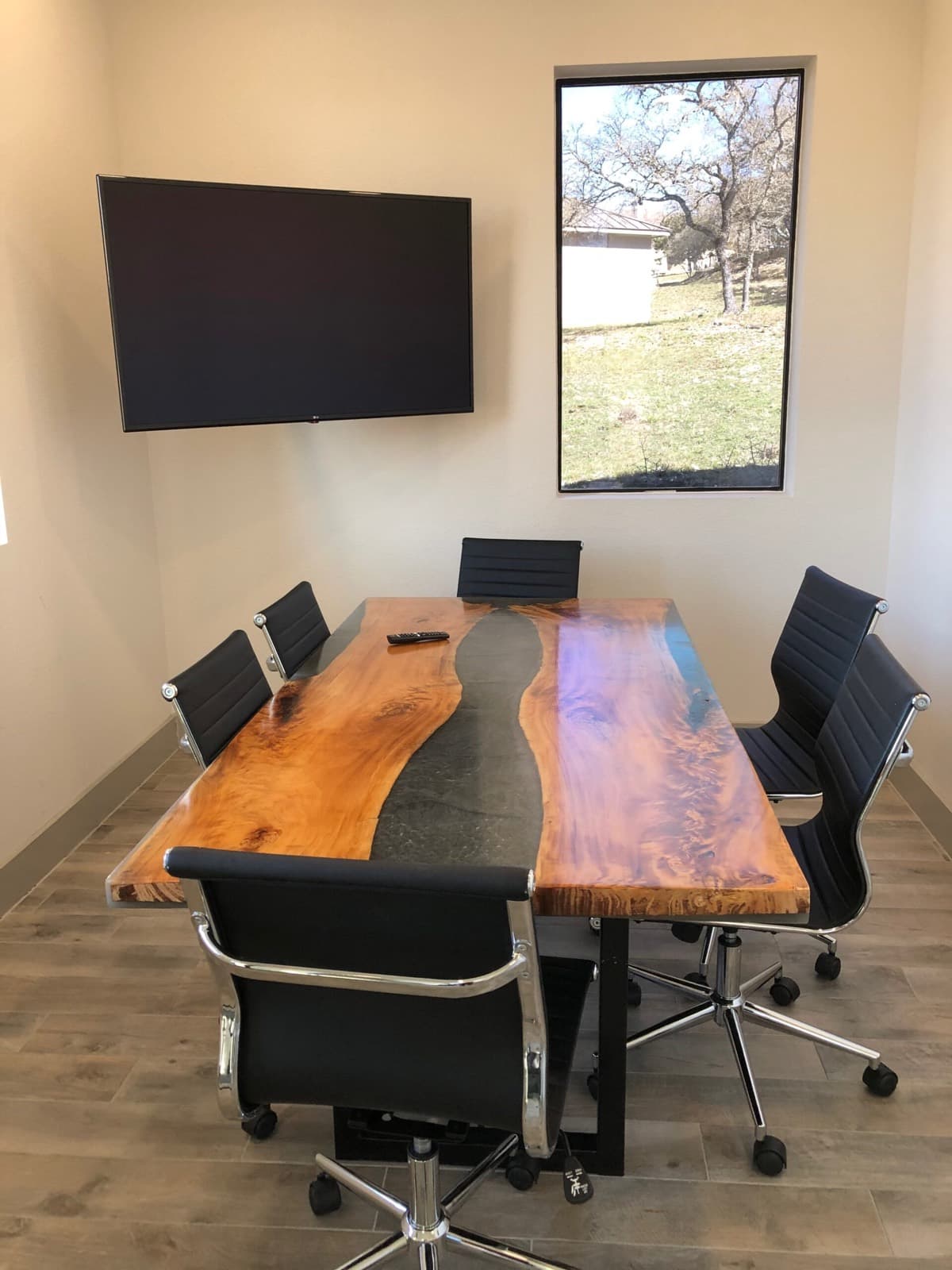 Conference room with a live-edge wood and resin table, black chairs, a wall-mounted screen, and a window onto live oaks.