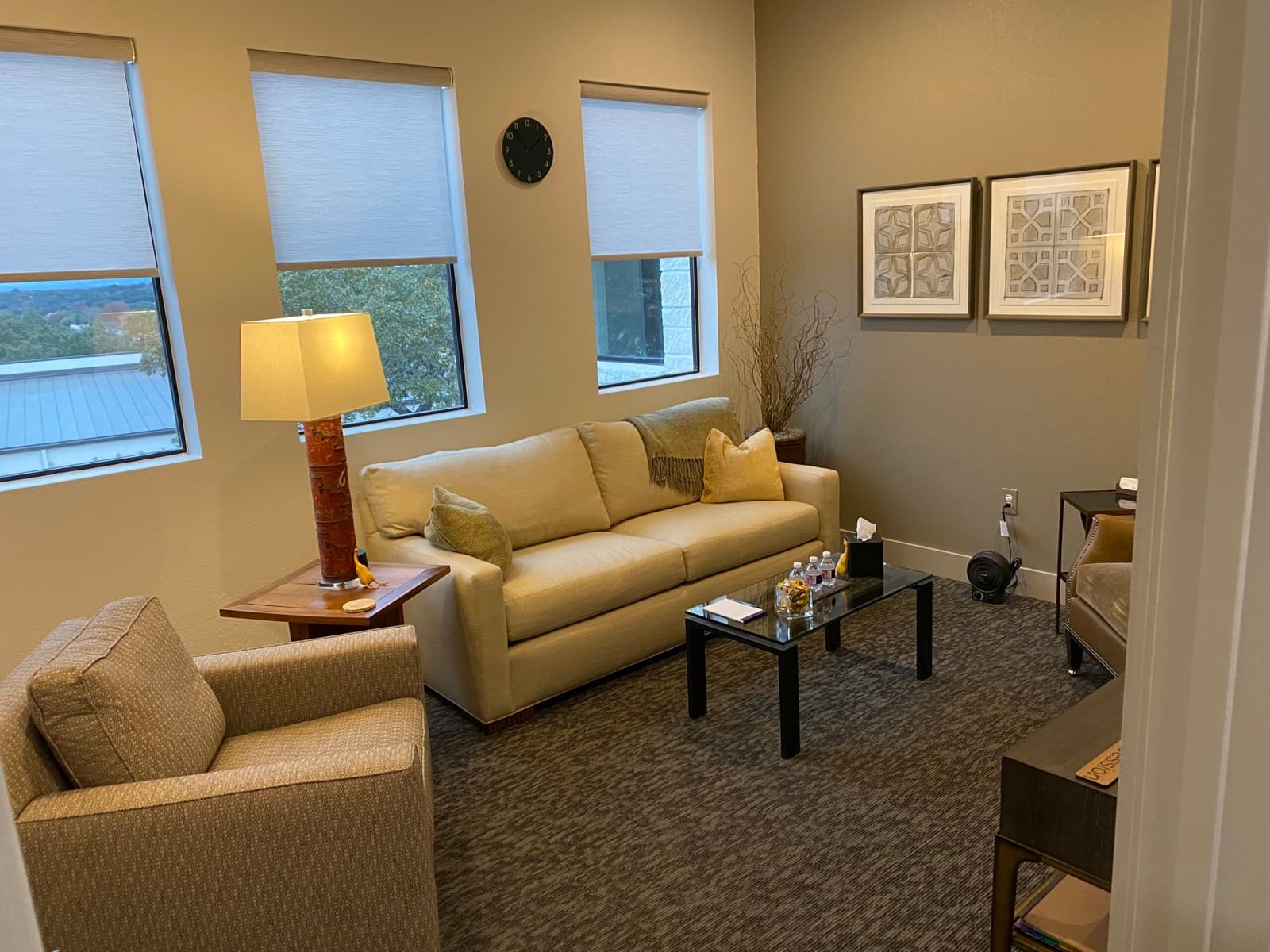Counseling room with a sofa, armchairs, and a glass coffee table beside three windows looking onto trees.