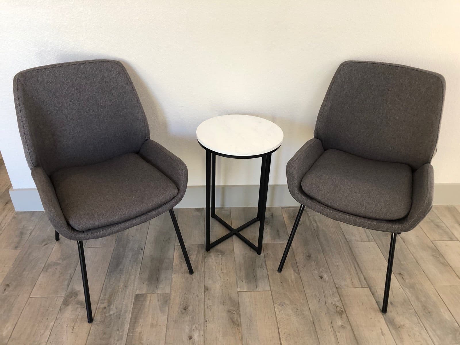 Two grey upholstered armchairs either side of a small round marble-topped table, against a white wall on wood-effect flooring.